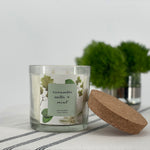 Candle with a label and cork lid on a white surface with greenery in the background