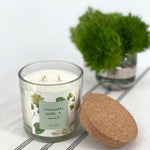 Candle with a label and a cork lid on a white surface with a plant in the background