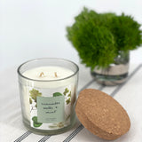Candle with a label and a cork lid on a white surface with a plant in the background