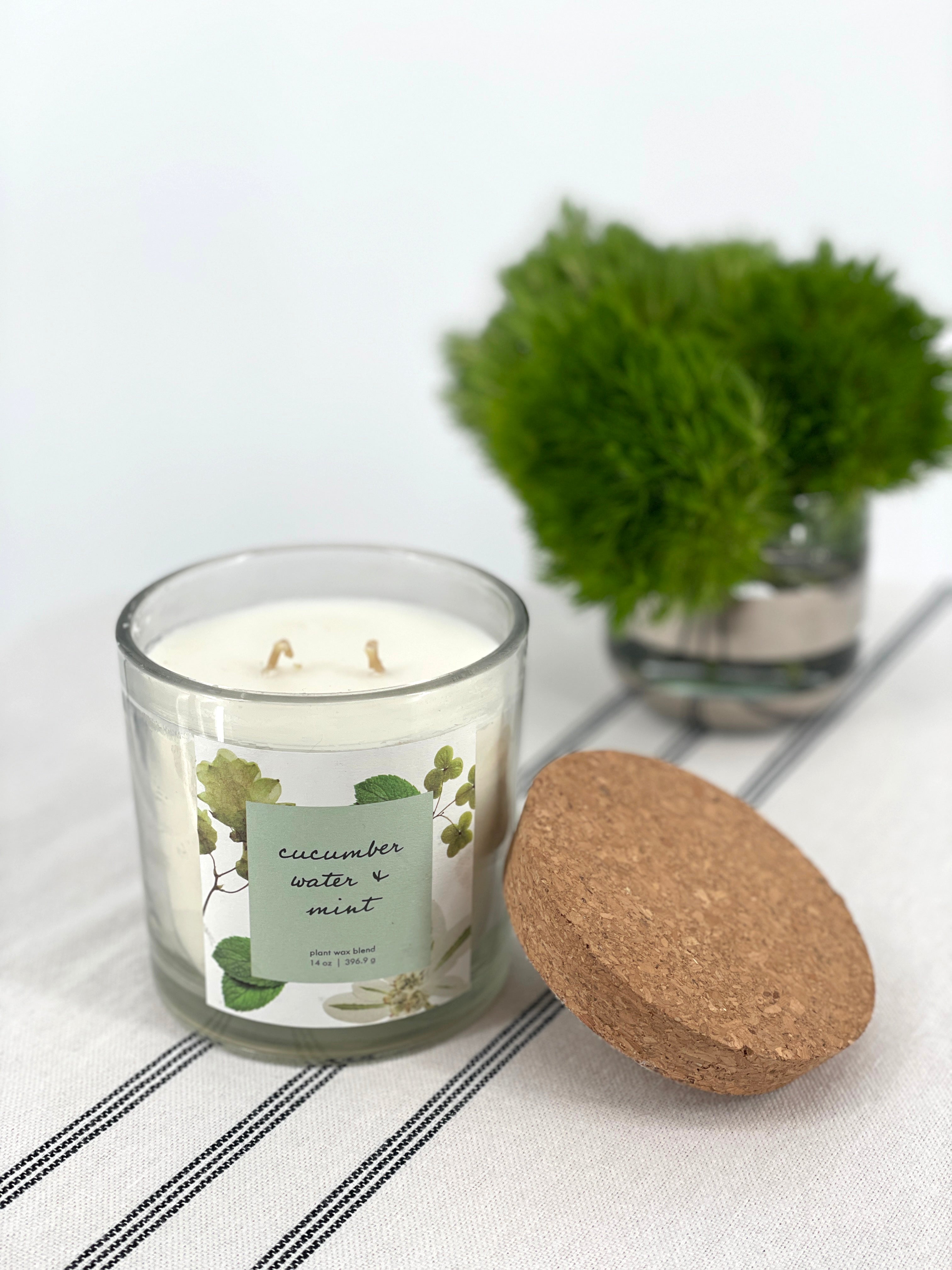 Candle with a label and a cork lid on a white surface with a plant in the background